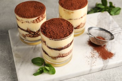 Photo of Delicious tiramisu in glasses, mint and sieve with cocoa powder on grey textured table, closeup