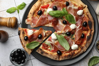 Tasty pizza with cured ham, olives, mozzarella cheese, sun-dried tomato and basil on light tiled table, flat lay