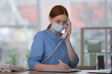 Photo of Receptionist with protective mask talking on phone at countertop in hospital