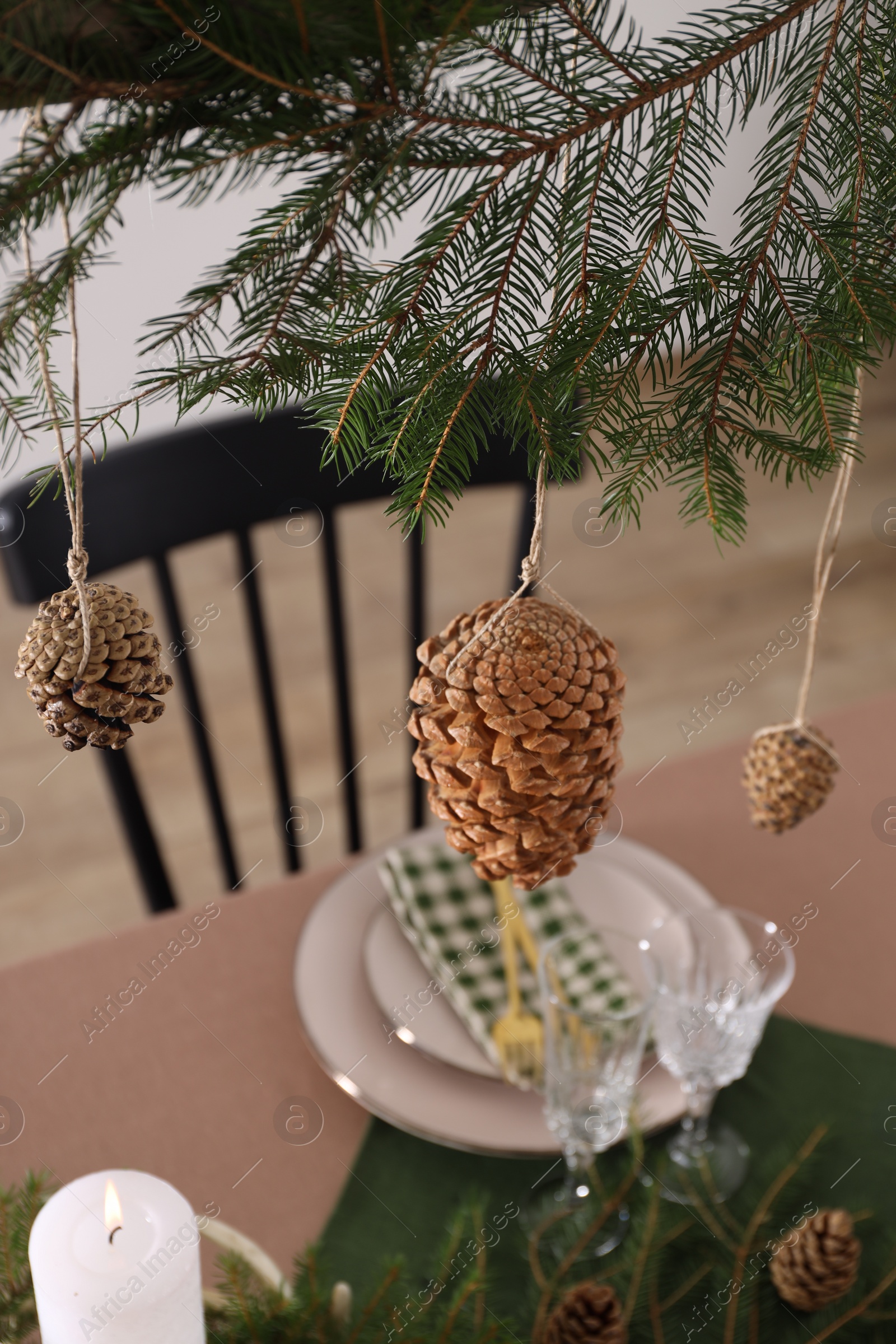 Photo of Christmas celebration. Cones hanging from fir tree branch over table with burning candle and tableware