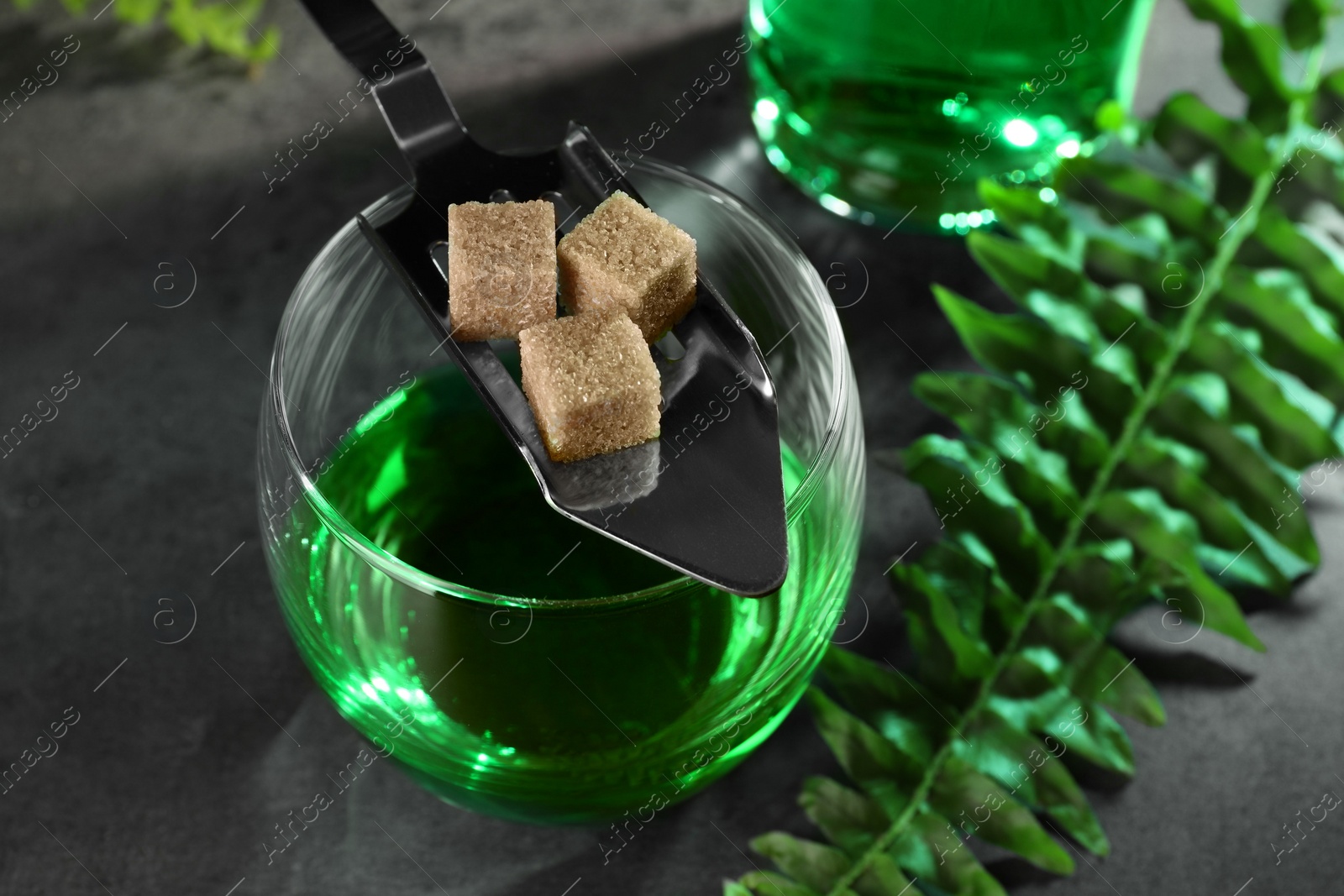 Photo of Absinthe in glass, spoon and brown sugar cubes on gray table, closeup. Alcoholic drink