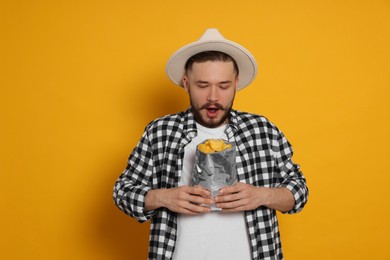 Emotional young man with bag of tasty potato chips on yellow background