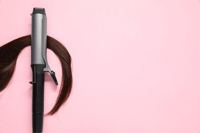 Photo of Curling iron with brown hair lock on pink background, top view. Space for text