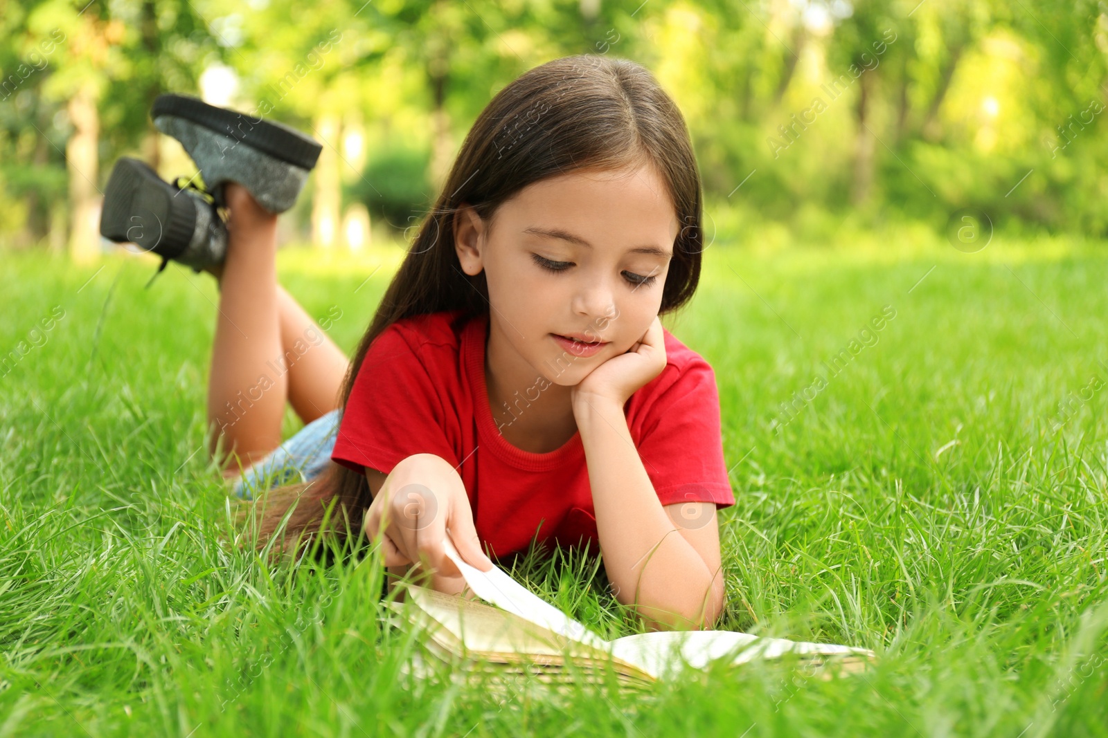 Photo of Cute little girl reading book on green grass in park