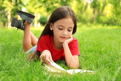 Photo of Cute little girl reading book on green grass in park