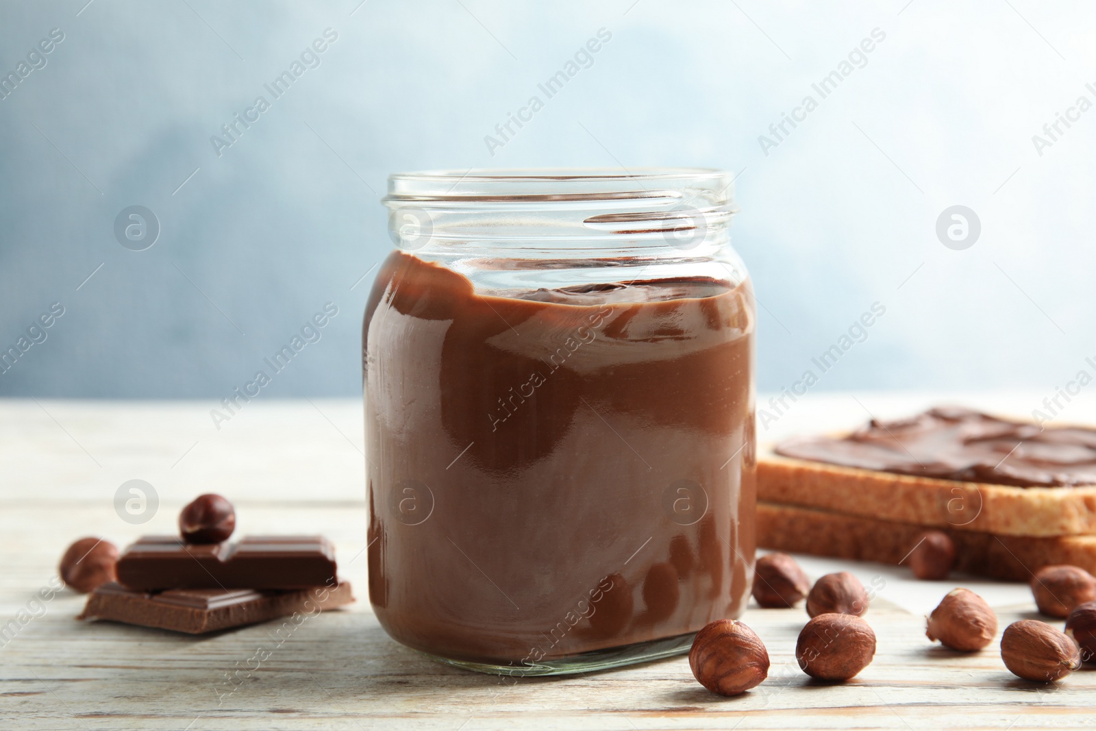 Photo of Glass jar with tasty chocolate cream and hazelnuts on wooden table against color background