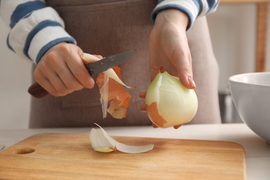Woman peeling fresh onion with knife at white table indoors, closeup