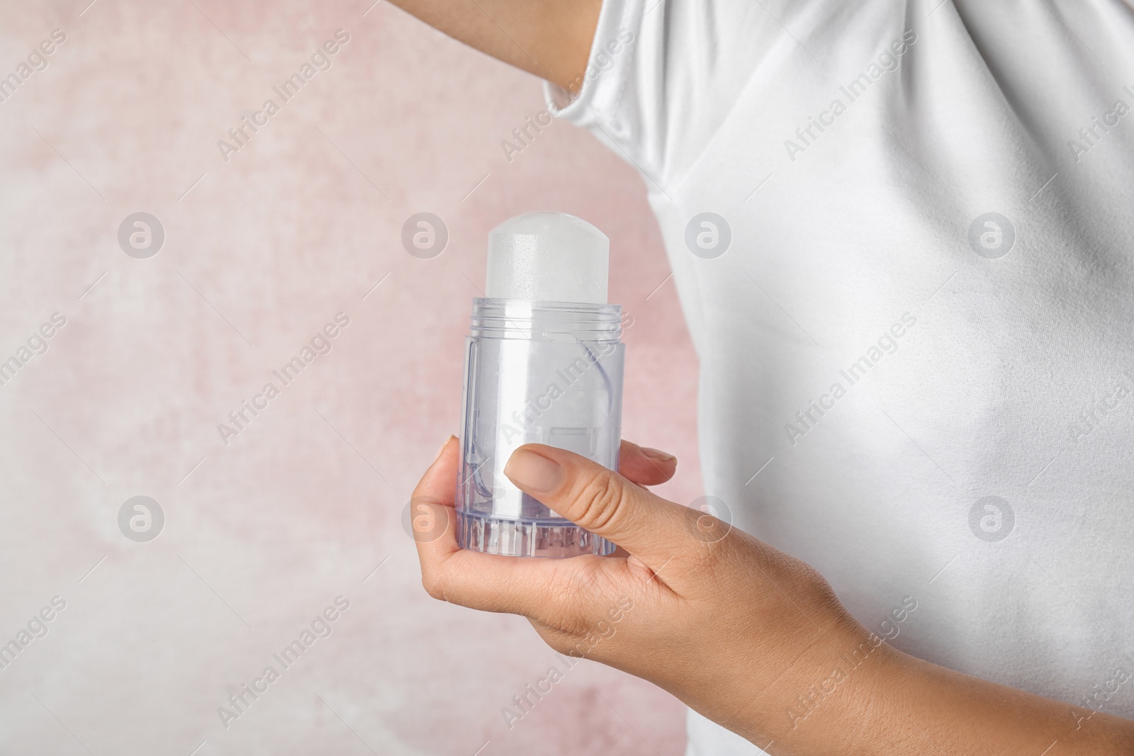 Photo of Young woman holding natural crystal alum deodorant near armpit on light pink background, closeup