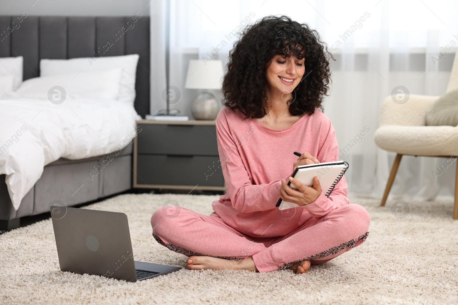 Photo of Beautiful young woman in stylish pyjama with laptop taking notes in notebook on floor at home