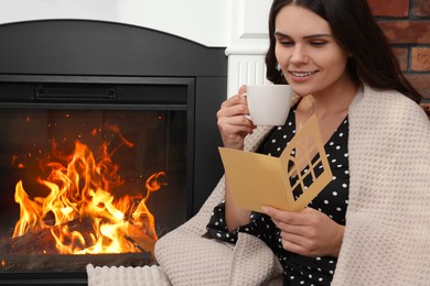 Young woman with greeting card and hot drink sitting near fireplace indoors