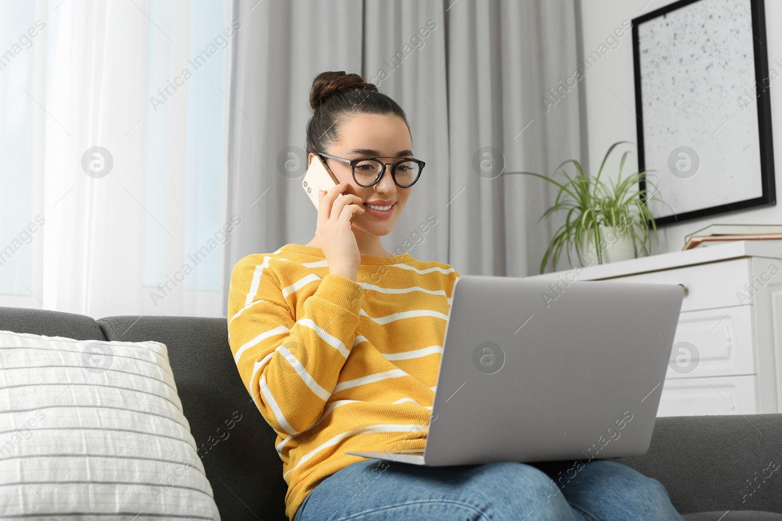 Photo of Home workplace. Happy woman with laptop talking on smartphone on sofa in room