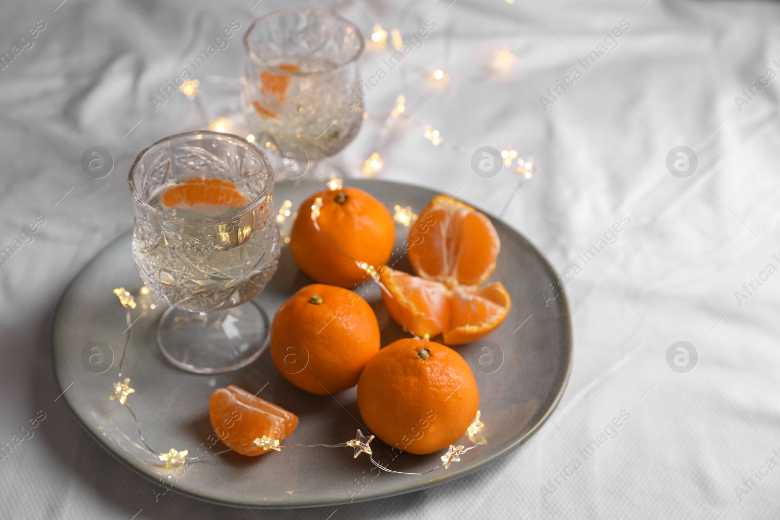 Photo of Delicious ripe tangerines, glasses of sparkling wine and fairy lights on white bedsheet. Space for text