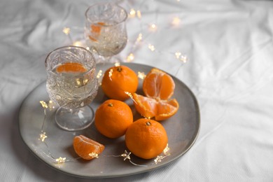 Photo of Delicious ripe tangerines, glasses of sparkling wine and fairy lights on white bedsheet. Space for text
