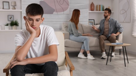 Unhappy teenage boy sitting in armchair while his parents arguing on background. Problems at home