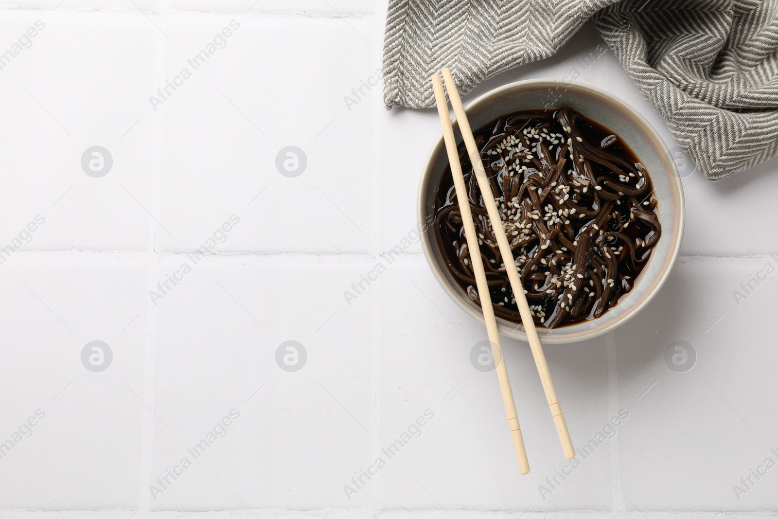 Photo of Tasty buckwheat noodles (soba) with sauce in bowl and chopsticks on white tiled table, top view. Space for text