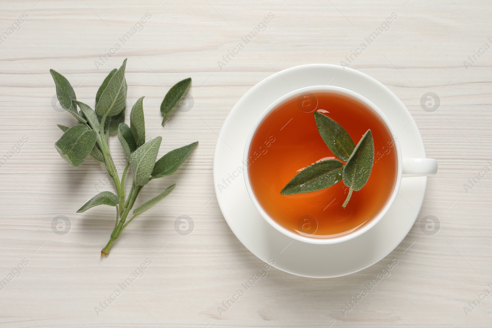 Photo of Cup of aromatic sage tea and fresh leaves on white wooden table, flat lay