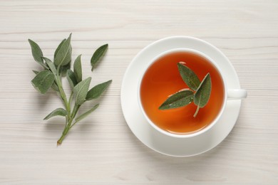 Cup of aromatic sage tea and fresh leaves on white wooden table, flat lay