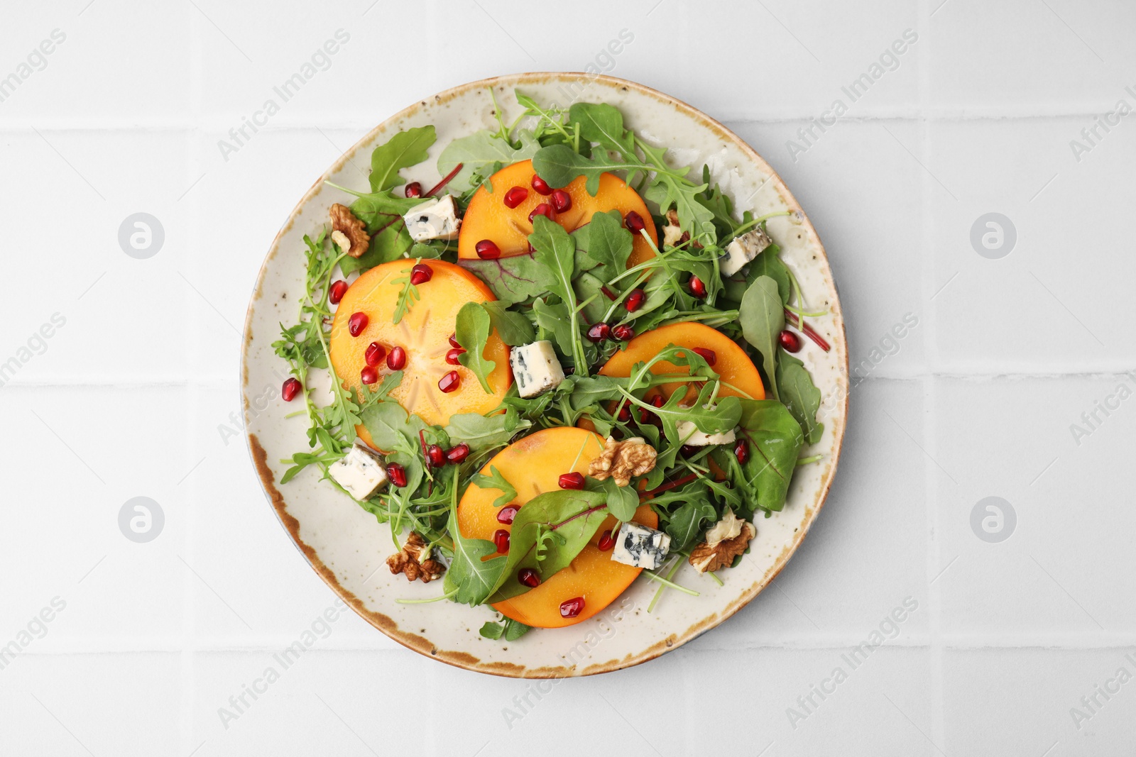 Photo of Tasty salad with persimmon, blue cheese, pomegranate and walnuts served on white tiled table, top view