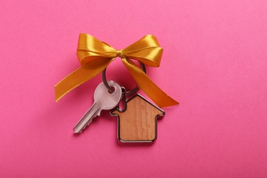 Key with trinket in shape of house and bow on pink background, top view. Housewarming party