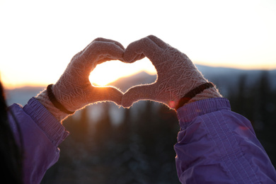 Woman making heart with her hands in mountains at sunset, closeup. Winter vacation