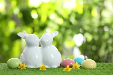 Cute ceramic Easter bunnies and dyed eggs on green grass against blurred background, space for text