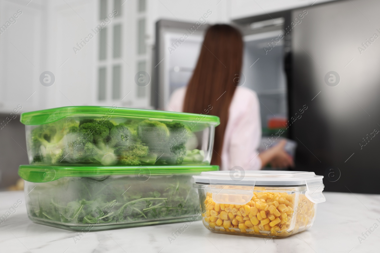 Photo of Glass containers with different fresh products on white marble table, selective focus. Woman near fridge in kitchen