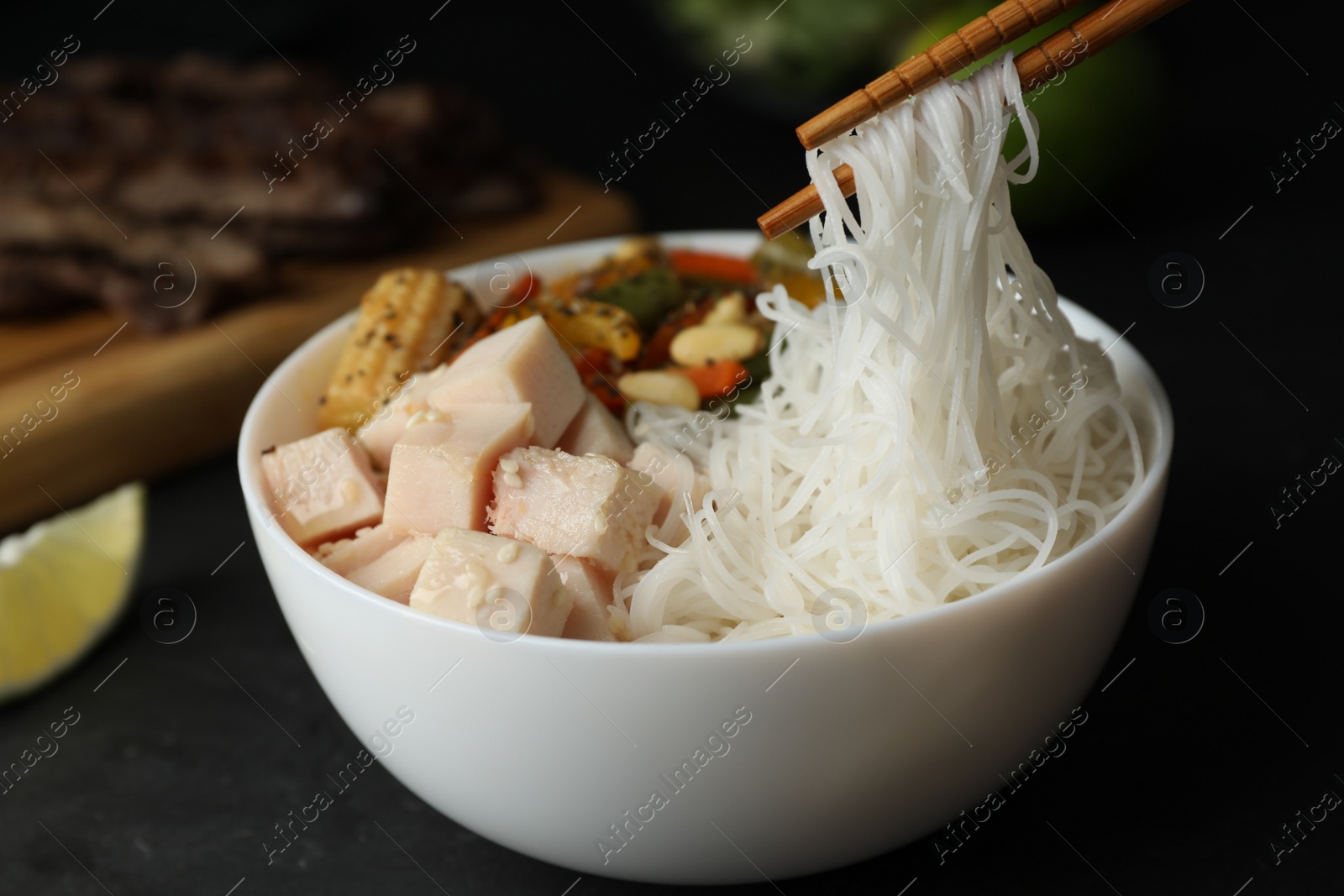 Photo of Chopsticks with tasty cooked rice noodles over bowl on black table, closeup