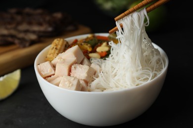Photo of Chopsticks with tasty cooked rice noodles over bowl on black table, closeup