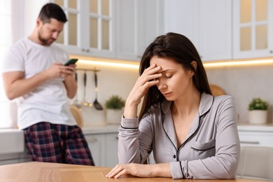 Photo of Offended wife sitting at table while her husband using smartphone in kitchen, selective focus. Relationship problems