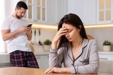 Offended wife sitting at table while her husband using smartphone in kitchen, selective focus. Relationship problems