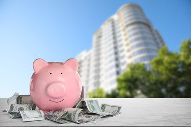 Image of Piggy bank and money on stone surface and blurred view of beautiful skyscraper, space for text. Mortgage concept