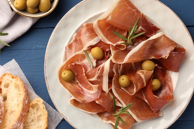 Photo of Slices of tasty cured ham, rosemary, bread and olives on blue wooden table, flat lay