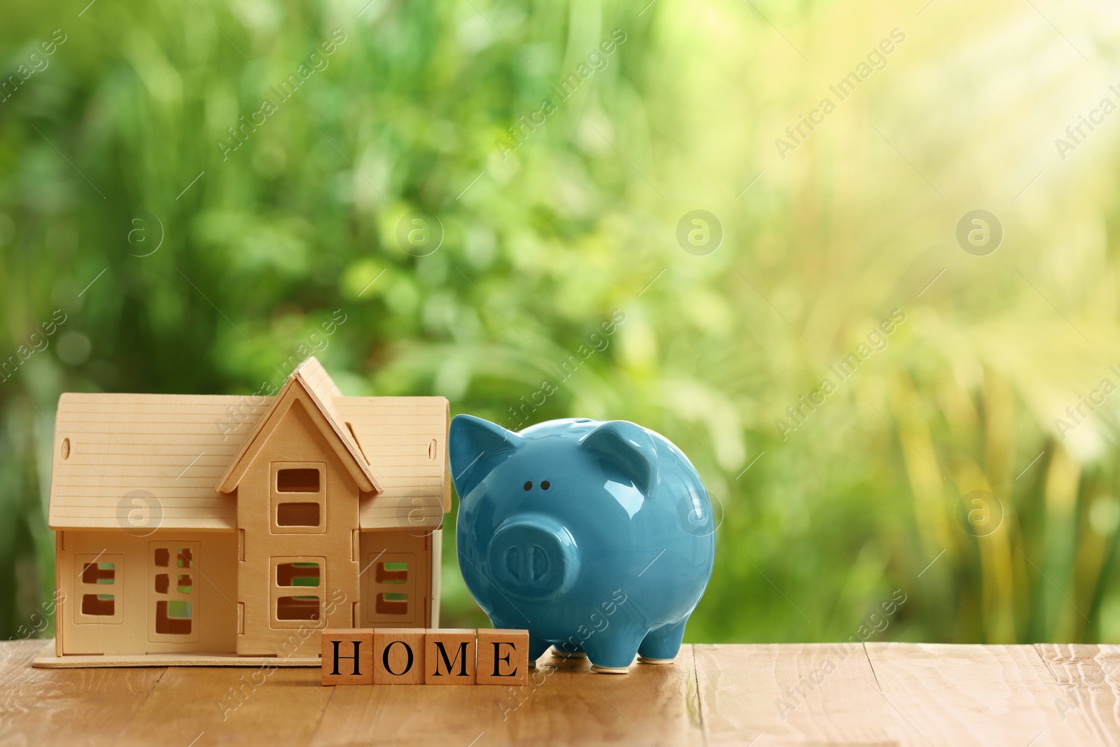 Photo of Piggy bank, house model and word Home made of cubes on wooden table outdoors. Space for text