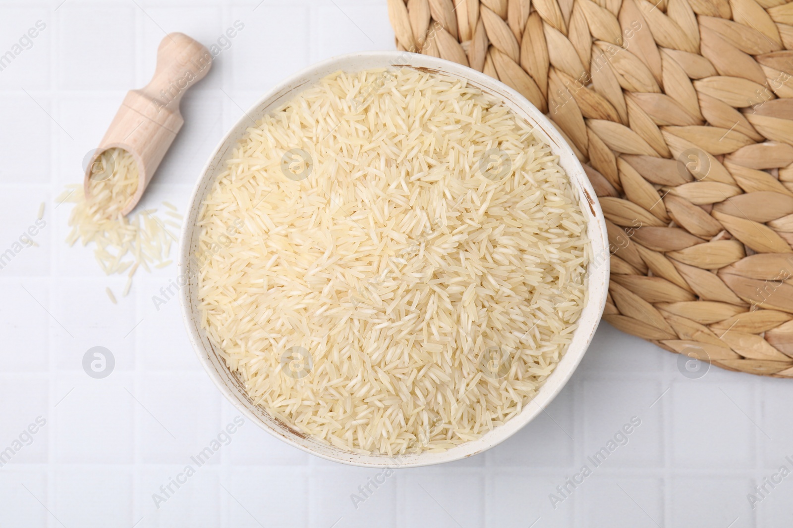 Photo of Bowl and scoop with raw rice on white tiled table, flat lay