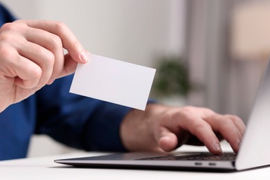 Man with laptop holding blank business card at white table indoors, closeup