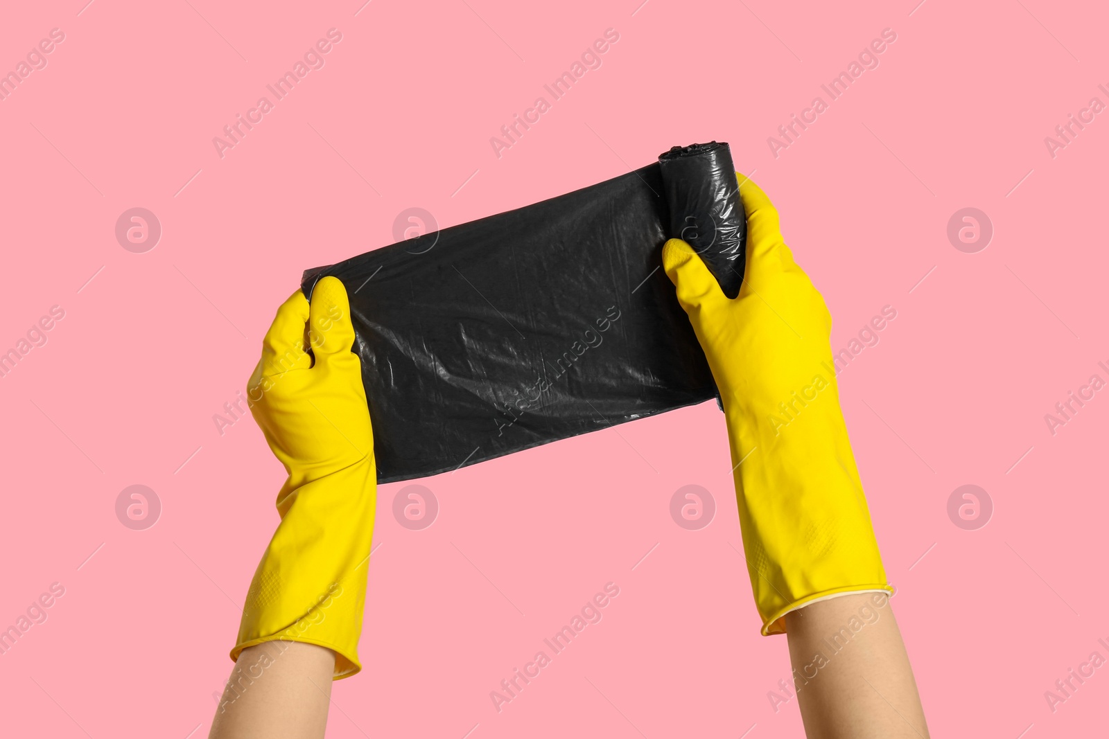 Photo of Janitor in rubber gloves holding roll of black garbage bags on pink background, closeup
