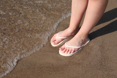 Woman in stylish white flip flops on sandy beach near sea, closeup