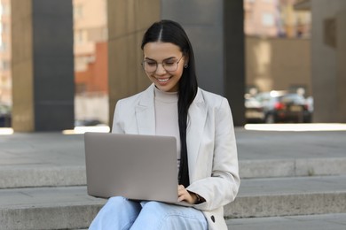 Photo of Happy young woman using modern laptop on stairs outdoors