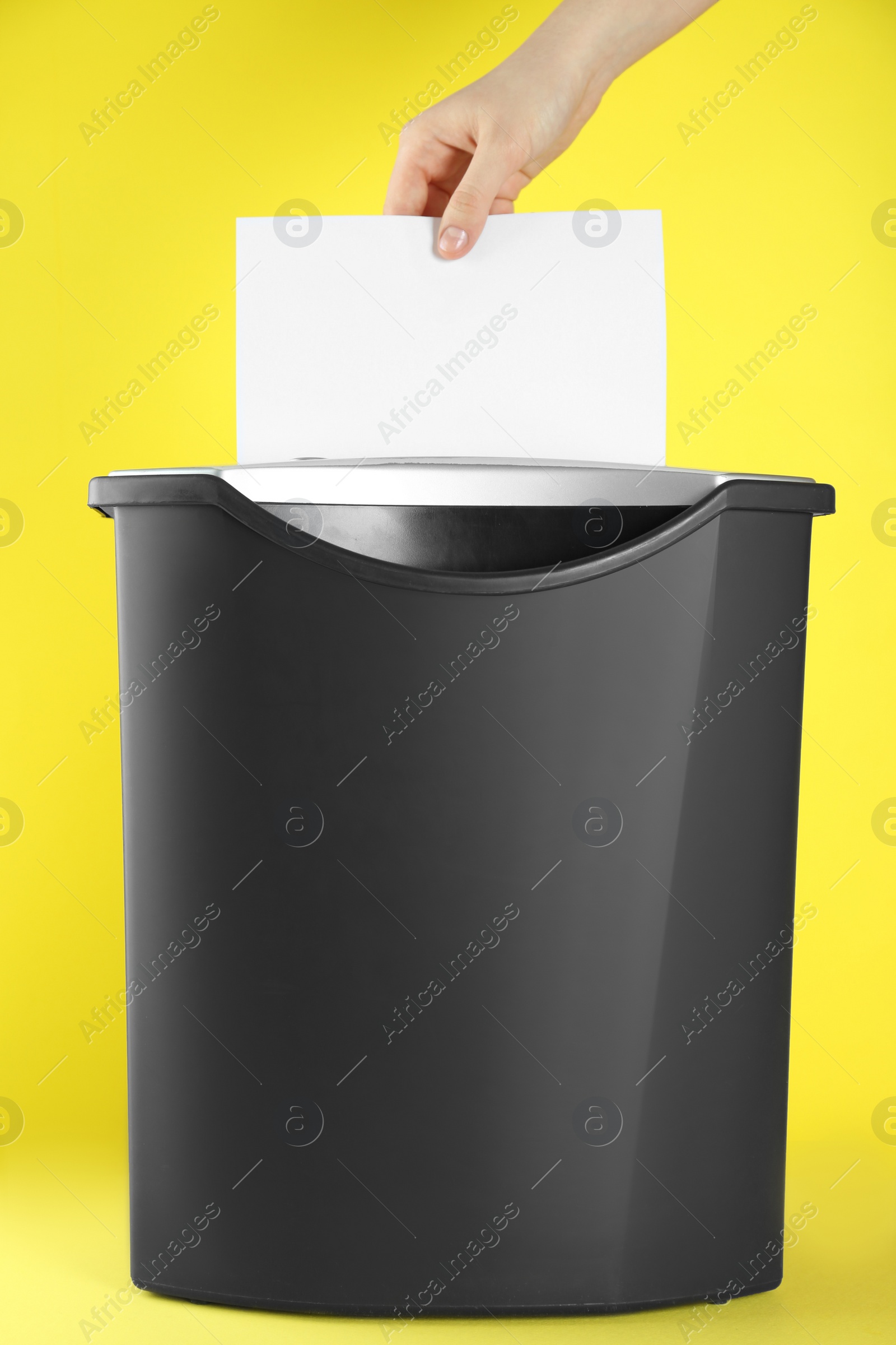 Photo of Woman destroying sheet of white paper with shredder on yellow background, closeup