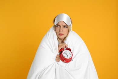 Photo of Sad young woman in sleeping mask wrapped with blanket holding alarm clock on yellow background