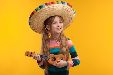 Cute girl in Mexican sombrero hat playing ukulele on orange background