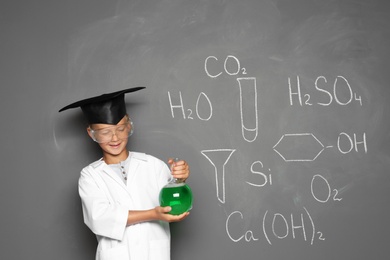 Little school child in laboratory uniform with flask of liquid and chemical formulas on grey background