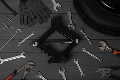 Car wheel, scissor jack, gloves and different tools on black wooden surface, flat lay