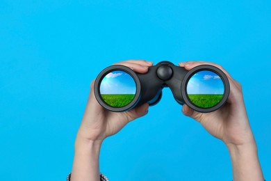 Image of Woman holding binoculars on light blue background, closeup. Green meadow reflecting in lenses