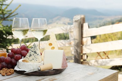 Photo of Different types of delicious cheeses, snacks and wine on wooden table against mountain landscape. Space for text