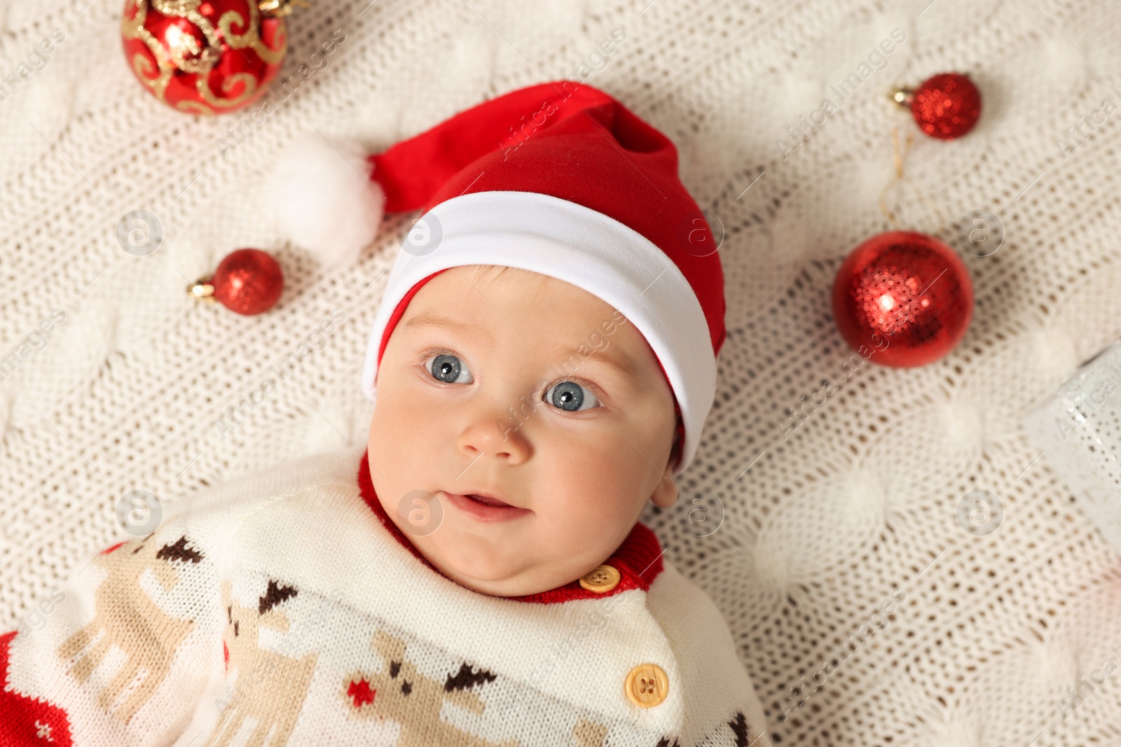 Photo of Cute little baby in Christmas outfit surrounded by  baubles on white knitted plaid, top view. Winter holiday