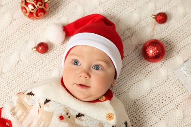Photo of Cute little baby in Christmas outfit surrounded by  baubles on white knitted plaid, top view. Winter holiday