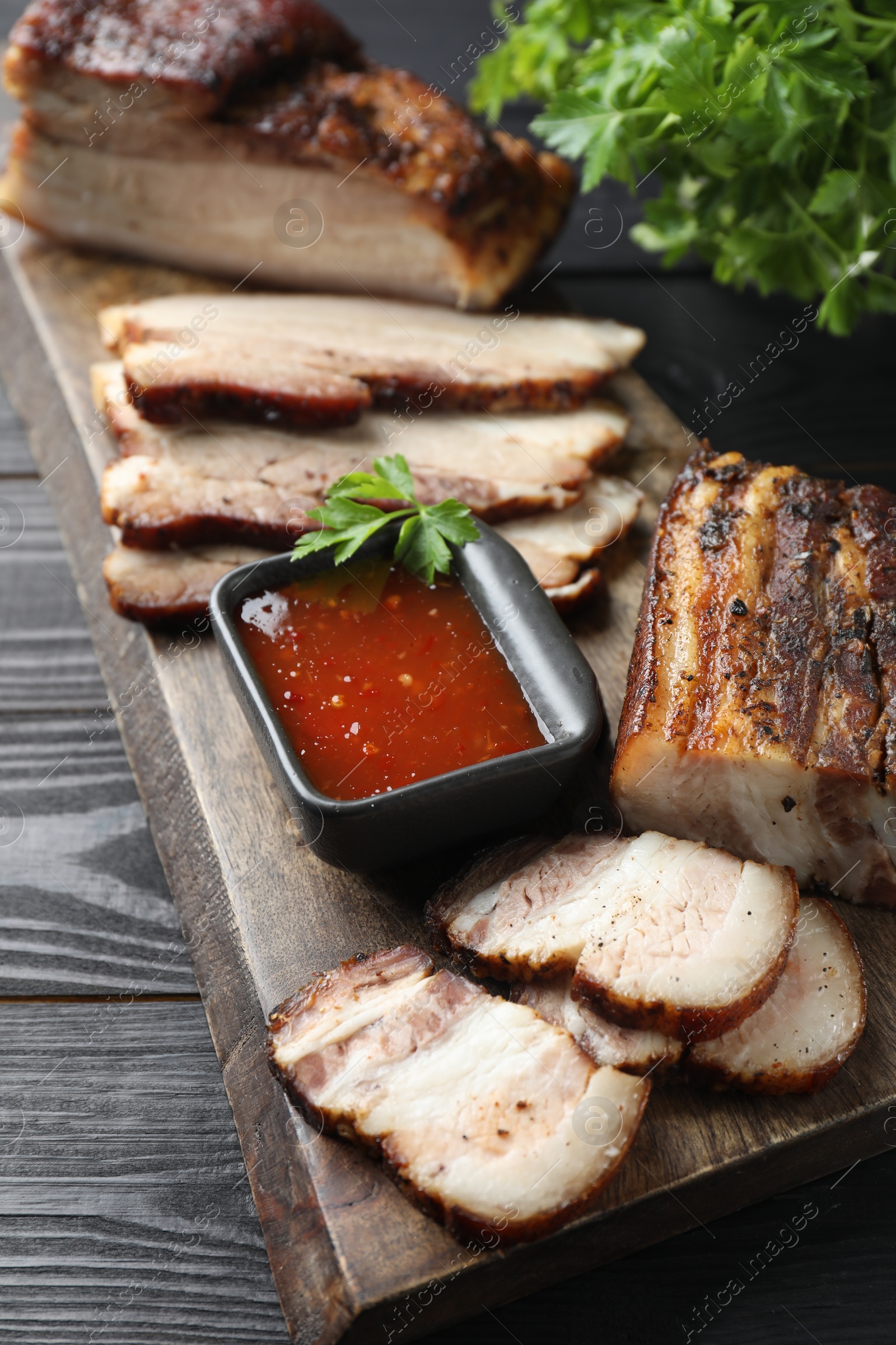 Photo of Pieces of baked pork belly served with sauce and parsley on black wooden table, closeup