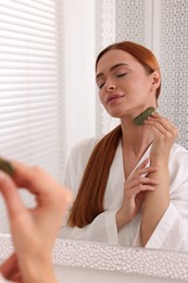 Young woman massaging her face with jade gua sha tool near mirror in bathroom