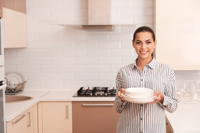 Photo of Beautiful young woman holding stack of clean dishes in kitchen